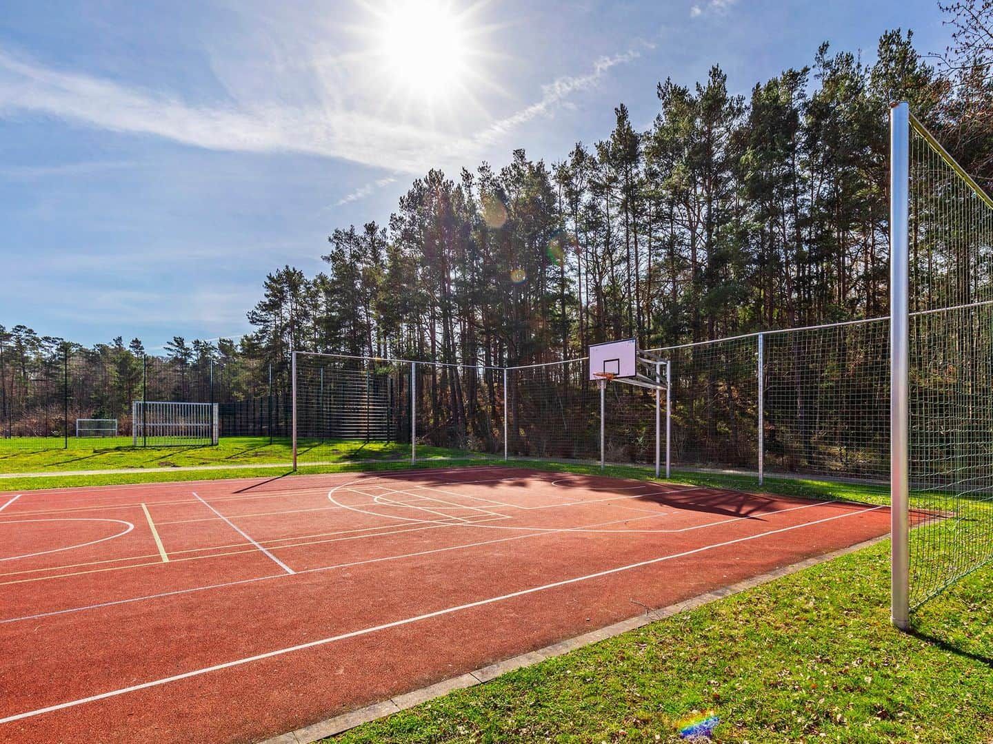 Una cancha de baloncesto al aire libre con superficie roja, rodeada por una cerca de metal y árboles.