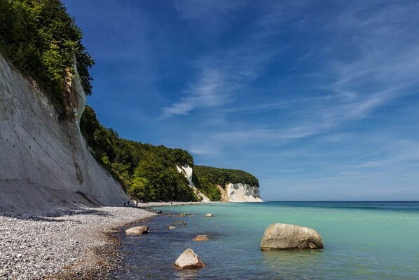 A coastal landscape with a rocky beach, clear blue waters, and a cliff with vegetation.