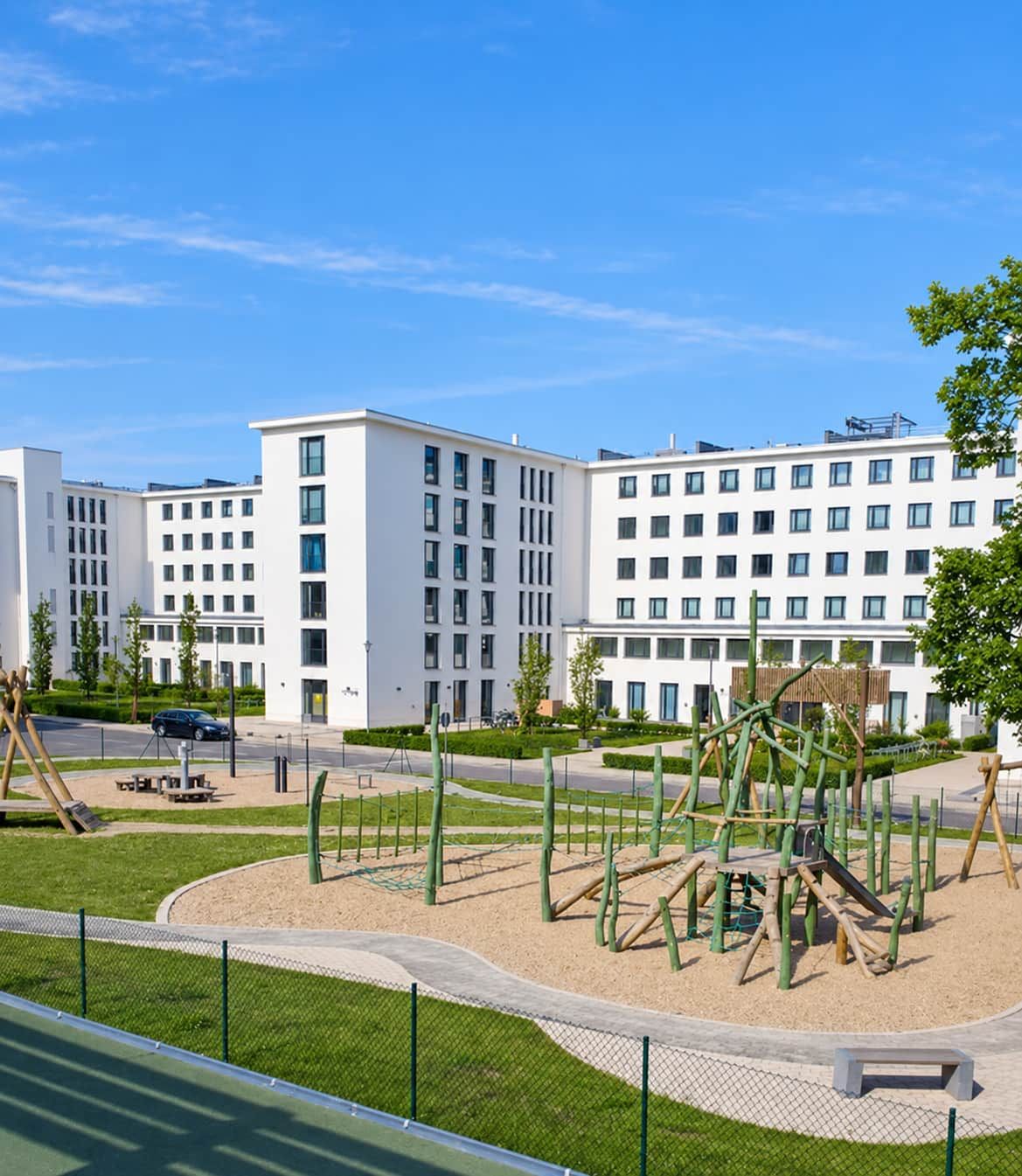 A playground with climbing structures and a fenced area, surrounded by a modern building complex with multiple floors and windows.