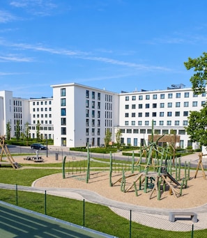 A playground with climbing structures and a fenced area, surrounded by a modern building complex with multiple floors and windows.