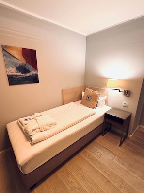 A single bed with a neatly folded bathrobe, a bedside table with a lamp, and a painting on the wall.