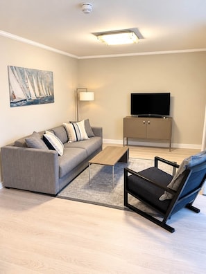 A living room with a sofa, coffee table, and armchair, a flat-screen TV on a cabinet, and a painting of sailboats on the wall.