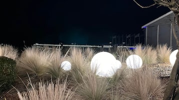 A nighttime outdoor area with tall grasses and illuminated spherical objects, a tree, and a building with flags in the background.