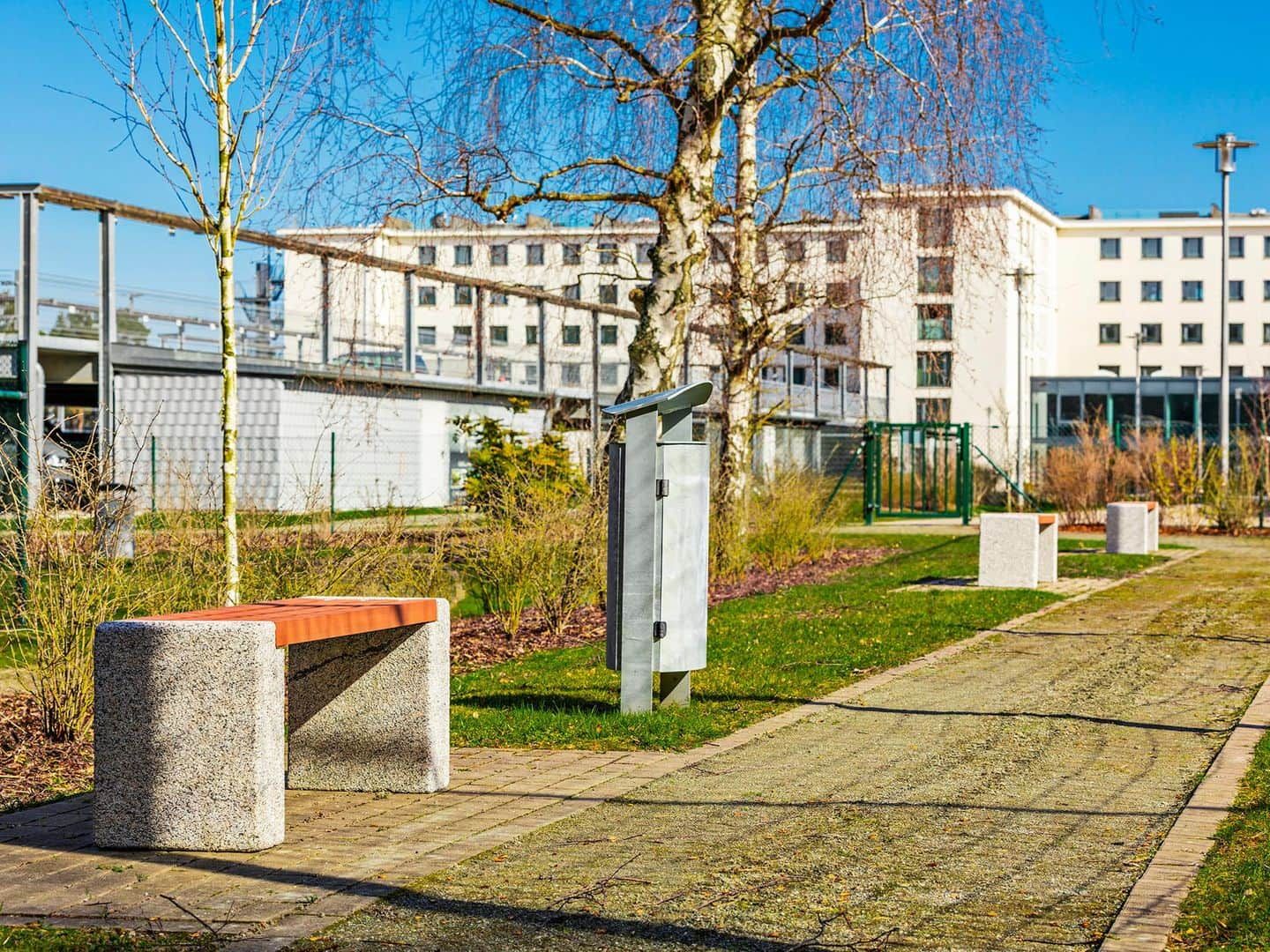 Ein Weg mit Bänken, einem Metallzaun und einem Gebäude im Hintergrund.