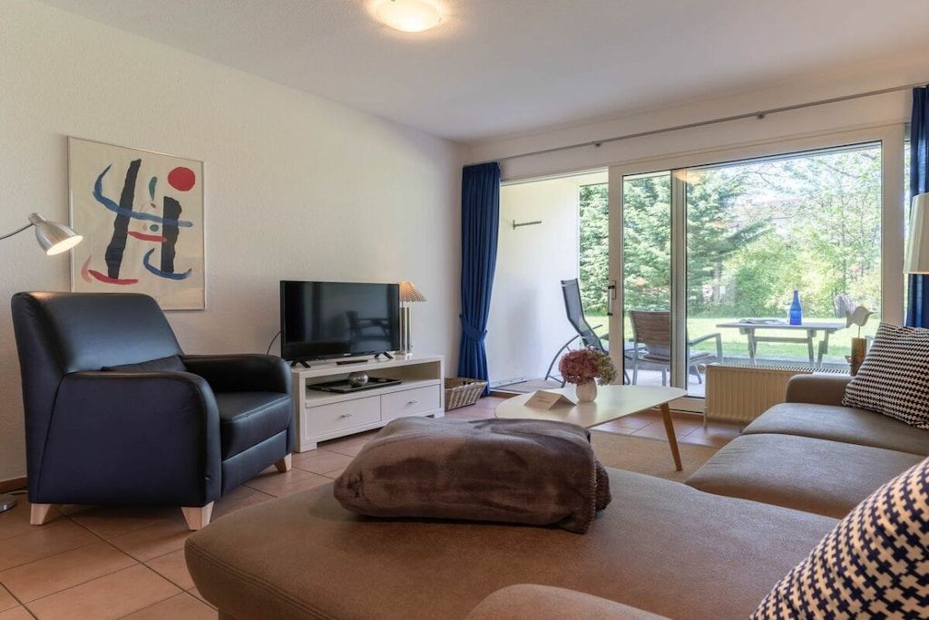 A modern living room with a flat-screen TV, a sofa, a chair, and a large window offering a view of the outdoors.
