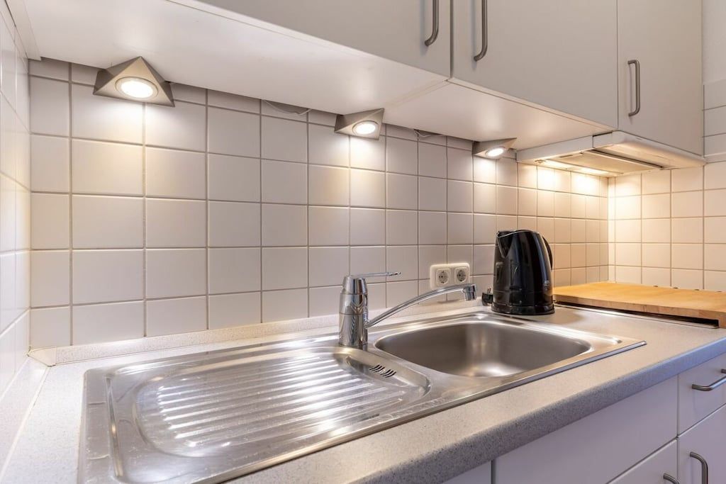 A modern kitchen with a stainless steel sink, a kettle, and white tiled walls.
