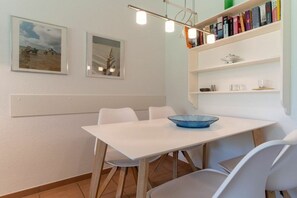 A dining area with a white table, chairs, and a blue bowl. There are framed pictures on the wall and a bookshelf with books. - Villa Anna II, Apartment 71 (St. Peter - Ording)