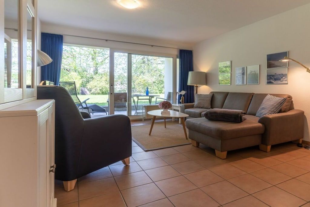 Sala de estar com sofá, poltrona e mesa de centro. Uma porta de vidro deslizante dá acesso a uma área externa.