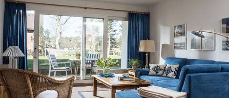 A living room with a blue sofa, a wicker chair, a coffee table, and a view through a glass door to an outdoor area.