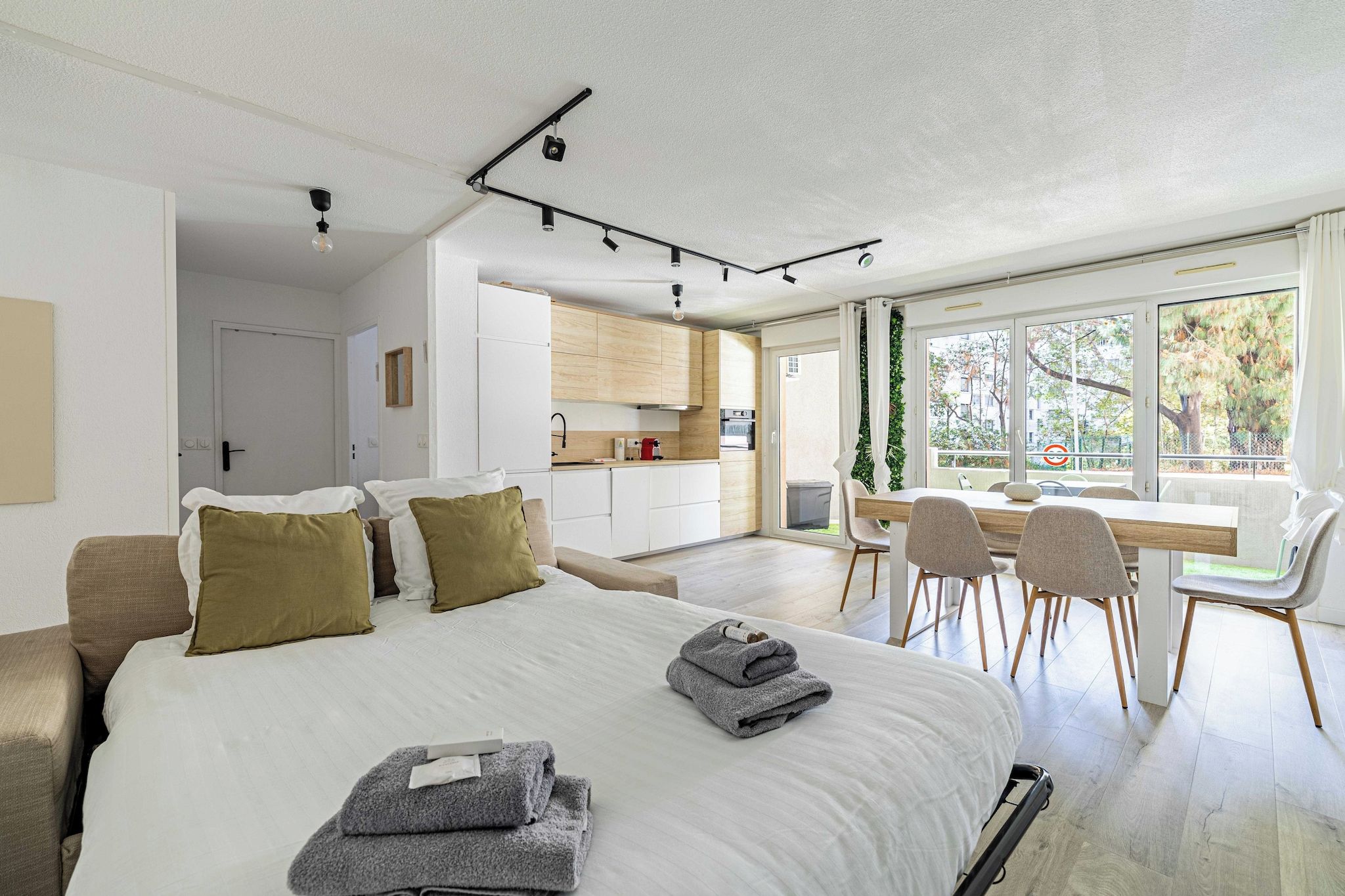 A modern bedroom with a large bed, a dining area with chairs, and a kitchen in the background.