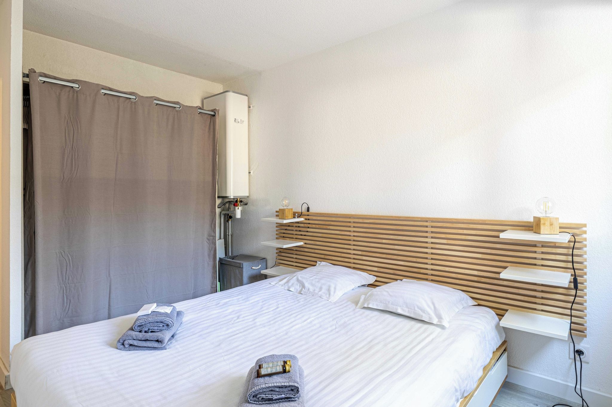 A hotel room with a bed, a wooden headboard, a bedside table, and a wall-mounted heater.