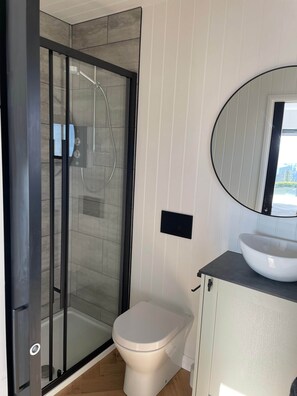 Un baño moderno con ducha de vidrio, inodoro blanco y espejo redondo sobre un lavamanos blanco.