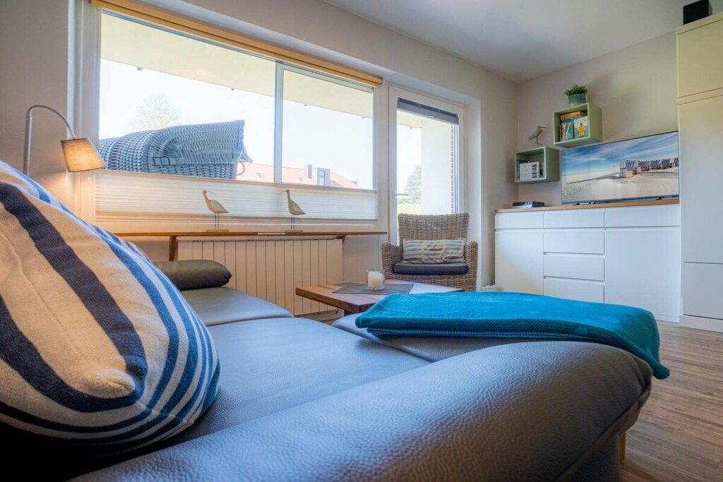 A cozy room with a sofa, a small table, a chair, a window with blinds, and a kitchen area with cabinets and a shelf.