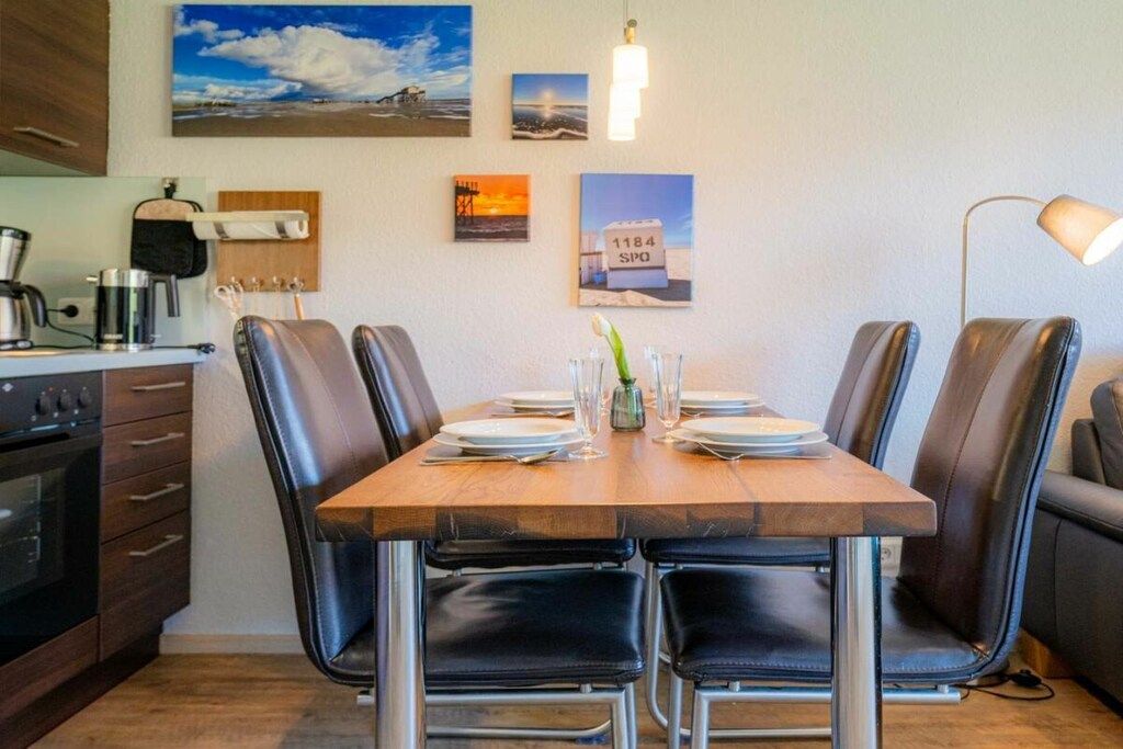Een eetgedeelte met een houten tafel voor vier personen, omringd door zwarte lederen stoelen. Links bevindt zich een keuken met een kookplaat en kastjes.