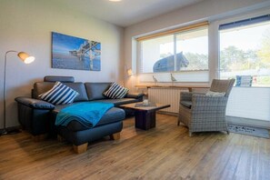 A modern living room with a sofa, ottoman, and wicker chair, a coffee table, a painting on the wall, and a window with blinds. - 2 in the Utlande house (ID 086) (St. Peter-Ording)