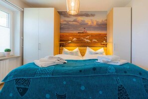 A bedroom with a bed, a headboard with a painting, and a window with blinds. - 2 in the Utlande house (ID 086) (St. Peter-Ording)