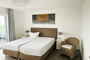 A double bed with white linens, two bedside tables with lamps, a wicker chair, and a painting on the wall. - Holiday apartment with 2 bedrooms (Wangerland)