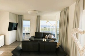 A modern living room with a black sofa, a flat-screen TV, and a large window offering a view of the city. - Holiday apartment with 2 bedrooms (Wangerland)