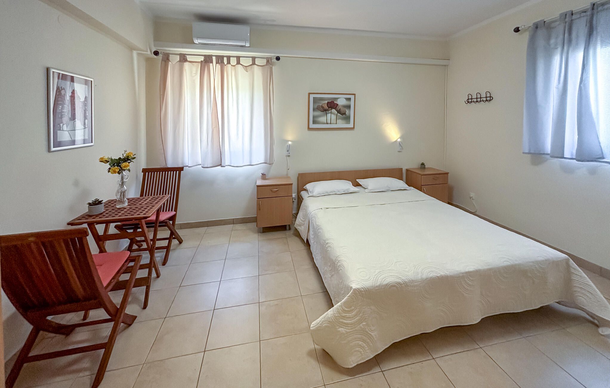 Een slaapkamer met een bed, nachtkastjes, een bureau met stoelen, een raam met gordijnen en een wandairconditioner.