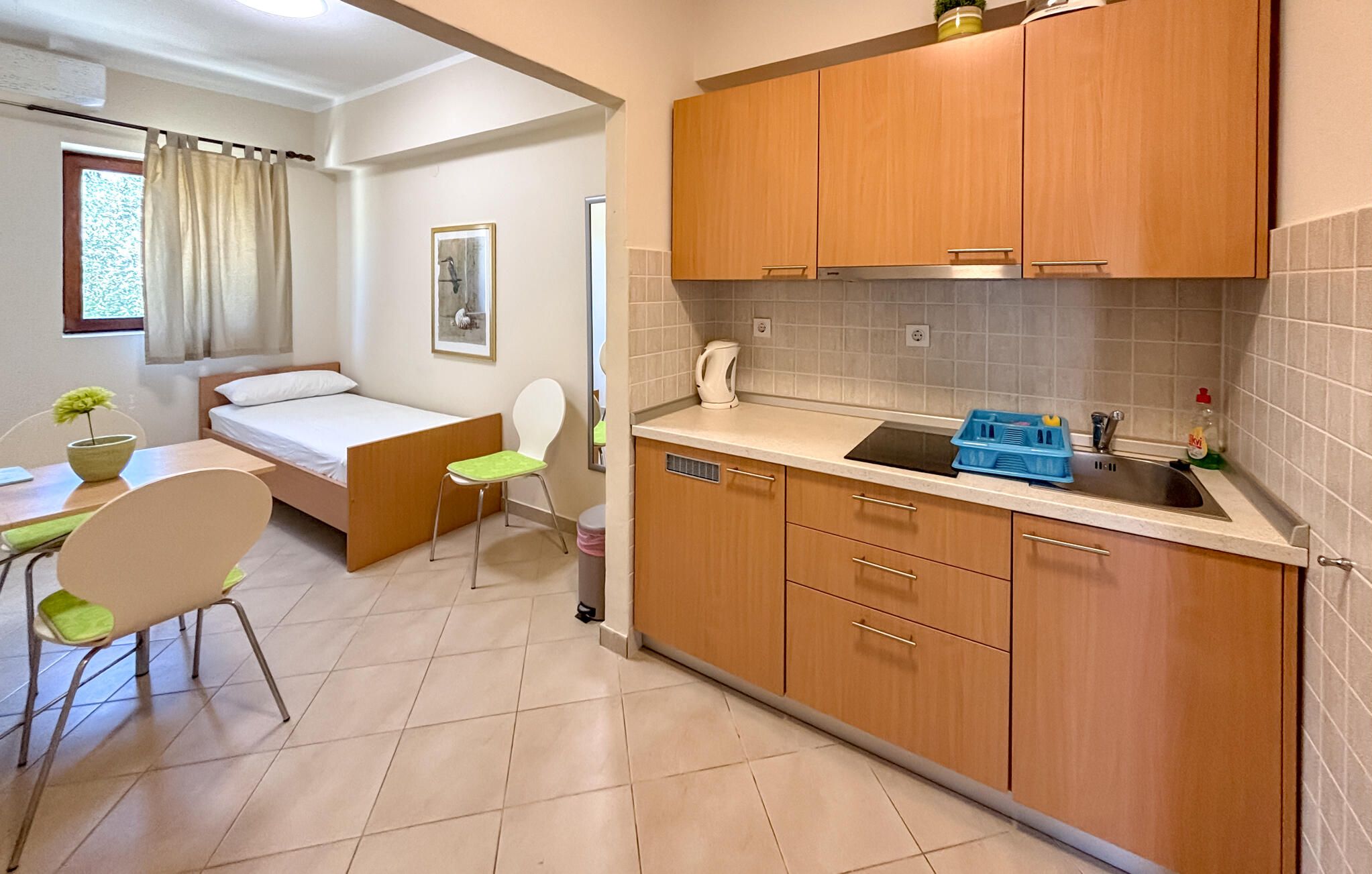 Cocina compacta con armarios de madera, un lavamanos y una encimera. En la habitación contigua hay una cama y una silla.
