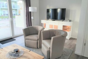 A modern living room with two beige armchairs, a flat-screen TV on a white cabinet, and a wooden coffee table with decorative items. - House No. 22, Deichgraf (Wangerland)