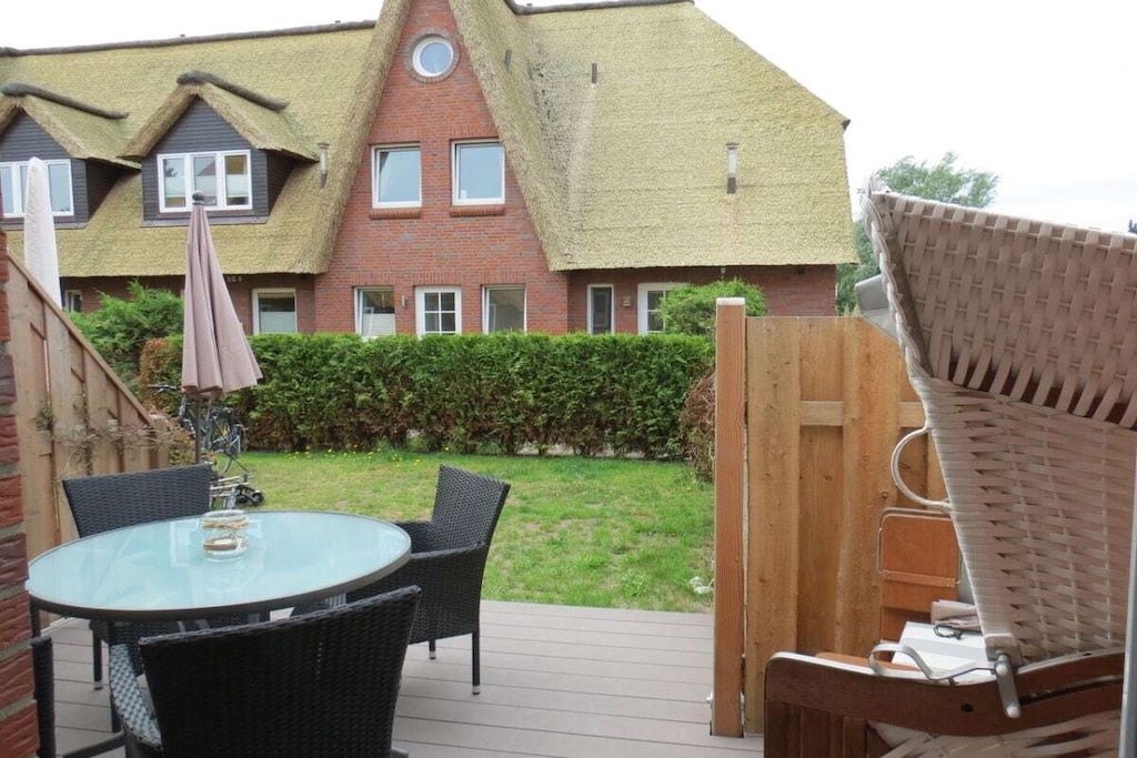 Une terrasse avec une table en verre et des chaises en osier, une clôture en bois et, au fond, une maison avec un toit de chaume.
