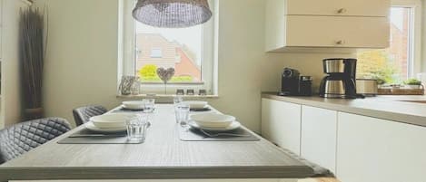 A dining area with a table set for four, a hanging light fixture, and a window with a view of a house.