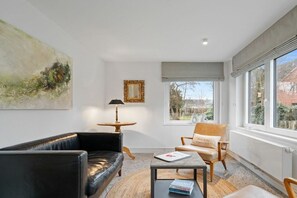 A modern living room with a black leather sofa, a wooden coffee table, and a beige armchair. There is a painting on the wall, a lamp, and a window with a view of the outside. - Sonnengold Apartment Sonnengold (St. Peter-Ording)