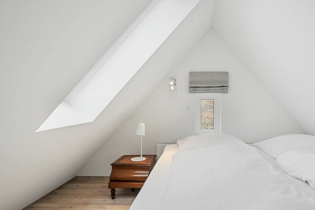 Ein Schlafzimmer mit schrägen Wänden, einem Nachttisch mit Lampe, einem Bett mit weißer Bettwäsche und einem Fenster mit Blick auf ein Gebäude.