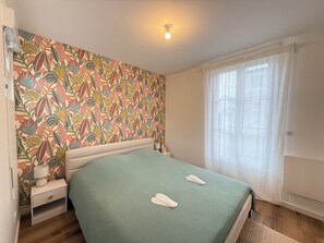 A bedroom with a floral wallpaper, a bed with a green bedspread, two white towels, a bedside lamp, and a window with sheer curtains. - Bright house for two, 200 m from the sea (Fouras)