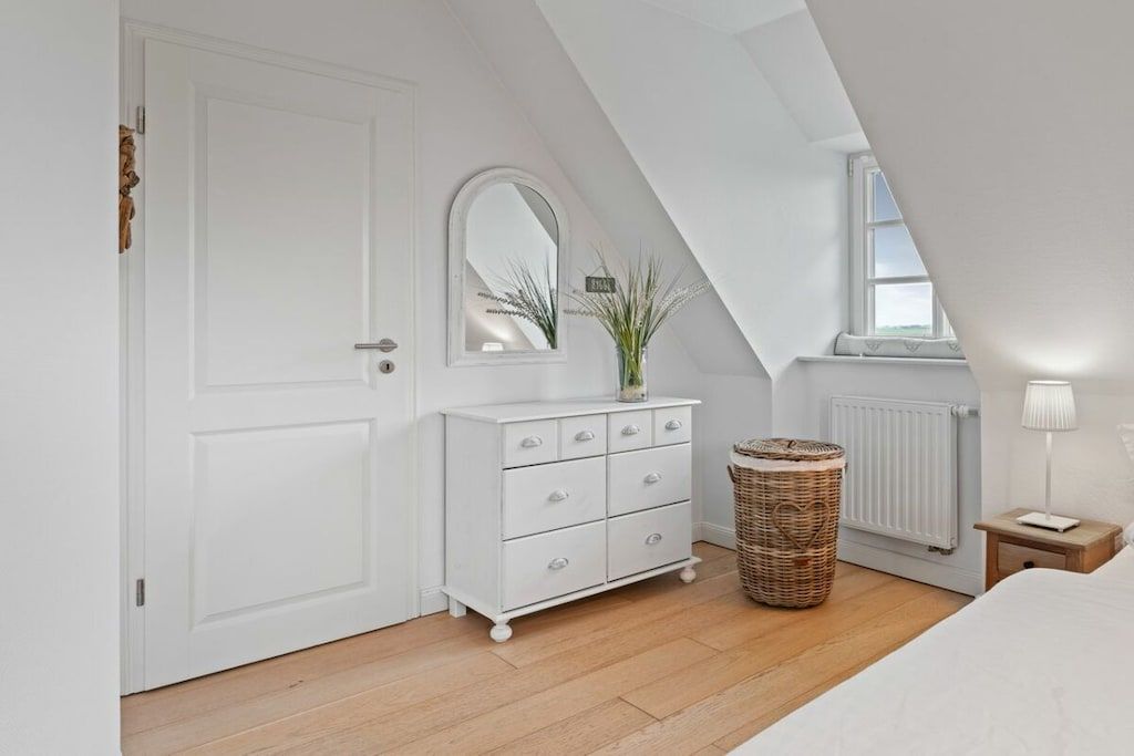 Een slaapkamer met een witte dressoir, een spiegel, een rieten mand, een radiator en een bedlamp.