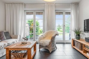 Ein Wohnzimmer mit einer Couch, einem Sessel, einem Couchtisch, einem Fernseher und einem großen Fenster mit Vorhängen.