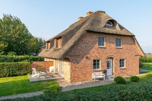 Ein Backsteinhaus mit einem Reetdach, einer Veranda mit Sitzgelegenheiten und einem gepflegten Rasen.