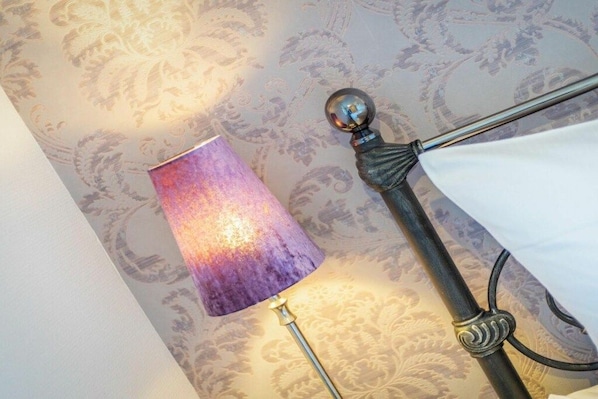 Ein Zimmer mit ornamentalem Wandpapier, einer Lampe mit Metallfuß und einem Bett mit dekorativer Kopfteil.