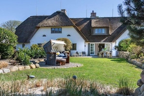 Ein Haus mit einem Reetdach, einem gepflegten Rasen und einer Terrasse mit Gartenmöbeln.