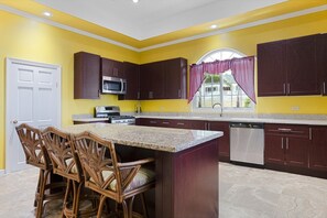 A kitchen with dark wooden cabinets, a granite countertop, a built-in microwave, and a window with curtains. - Hospitality Expert VSV TF 3BR - Chef, Pool, Beach (Montego Bay)