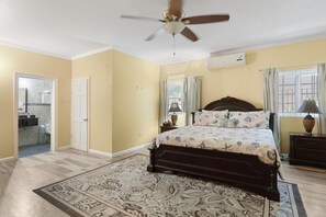 Ein Schlafzimmer mit Bett, Deckenventilator, Fenster mit Vorhängen und einem durchgehenden Türspalt sichtbaren Badezimmer.