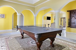 A billiards table in a room with yellow walls and white arches, a patterned carpet, and a television in the background. - Hospitality Expert VSV 6BR - Chef, Pool, Beach (Montego Bay)