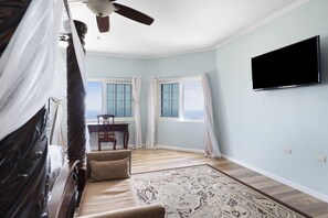 A room with a ceiling fan, a flat-screen TV mounted on the wall, a large window with curtains, a wooden desk, a cushioned sofa, and a patterned rug. - Hospitality Expert VSV GF 3BR - Chef, Pool, Beach (Montego Bay)