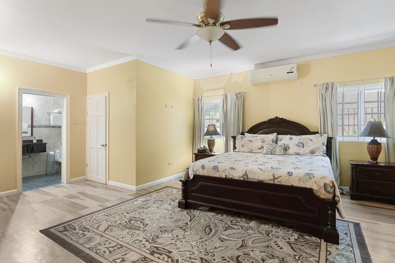 Une chambre à coucher avec un lit, un ventilateur de plafond, une fenêtre avec des rideaux et une salle de bain visible à travers une porte ouverte.