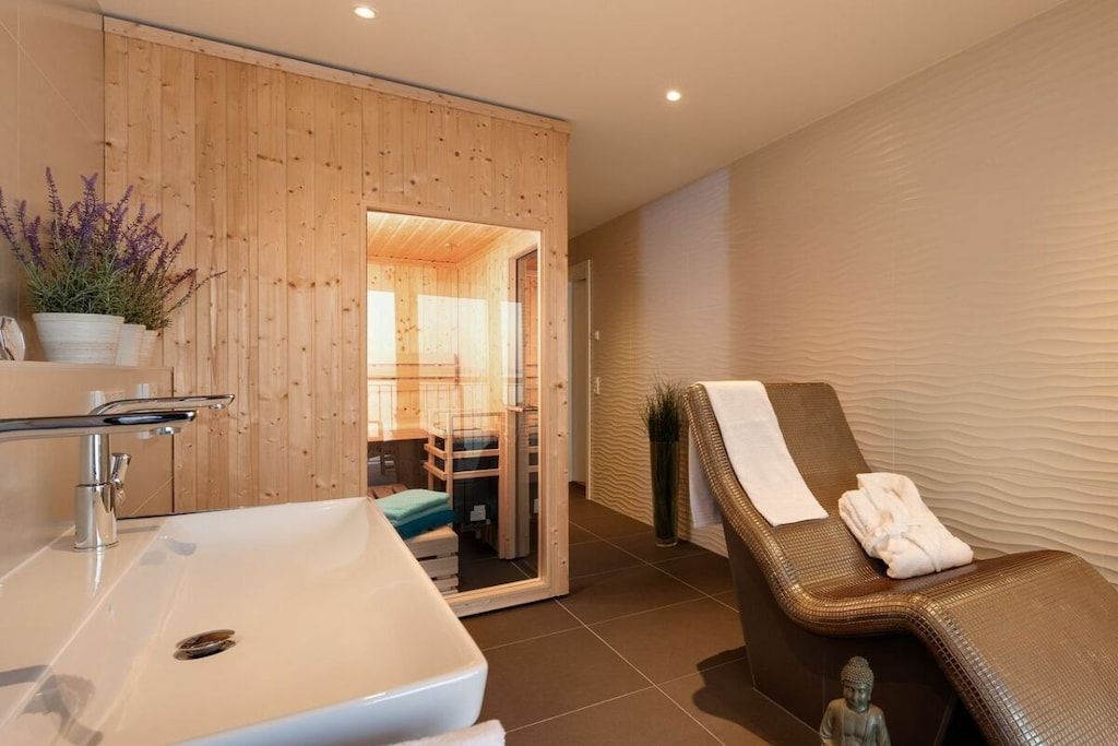A bathroom with a wooden wall, a sink, a mirror, and a relaxation area with a chair and towel.