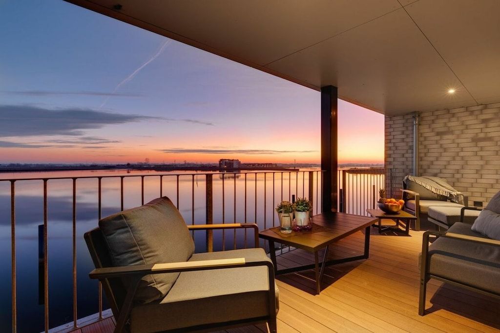 A balcony with a view of a sunset over a body of water, furnished with a table and chairs.