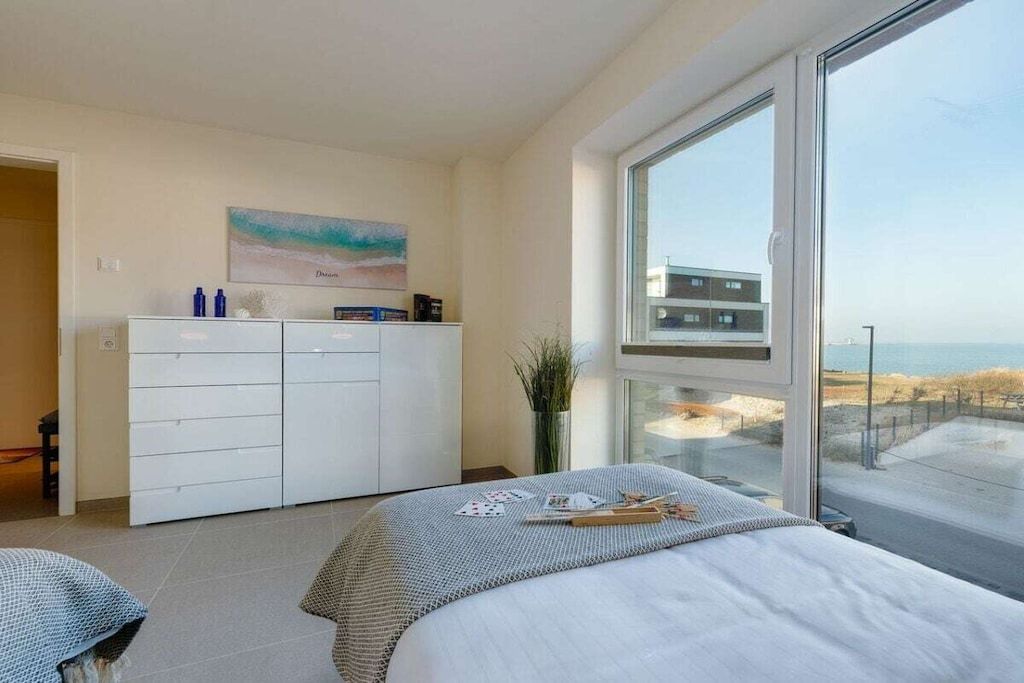 A bedroom with a bed, a dresser, a painting, and a window with a view of a building and beach.