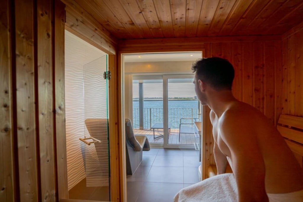 Interior de una cabaña de madera con sauna, puerta de cristal que da a un balcón y vista al mar.