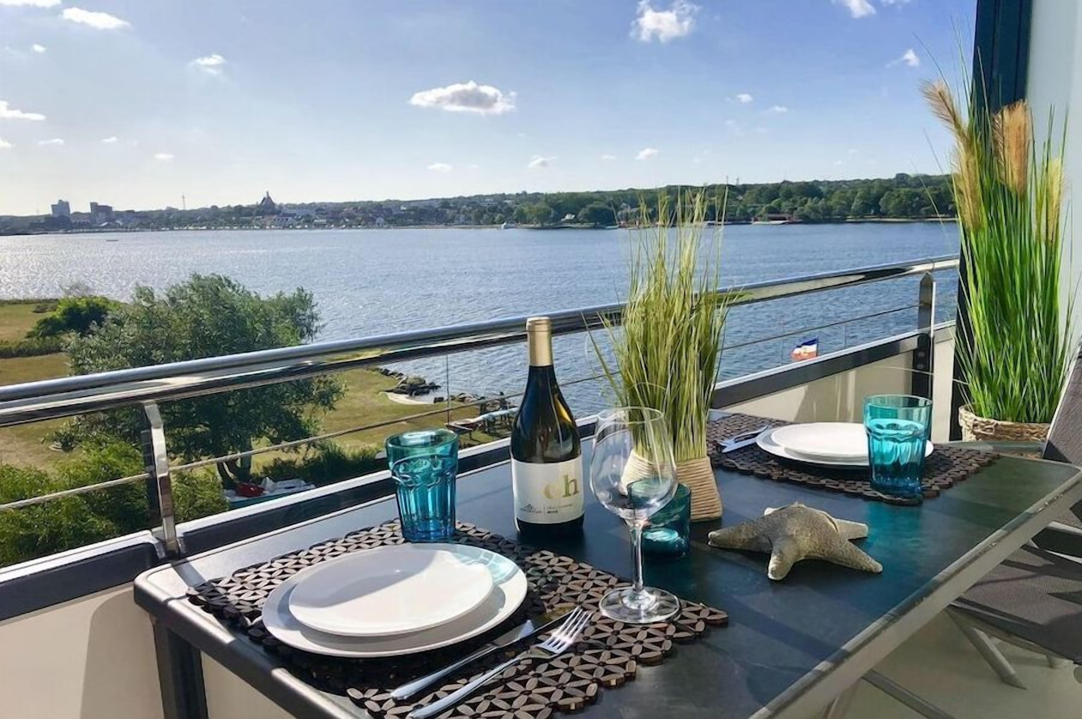A dining table with a bottle, glasses, and a starfish decoration, overlooking a body of water with a distant shoreline.