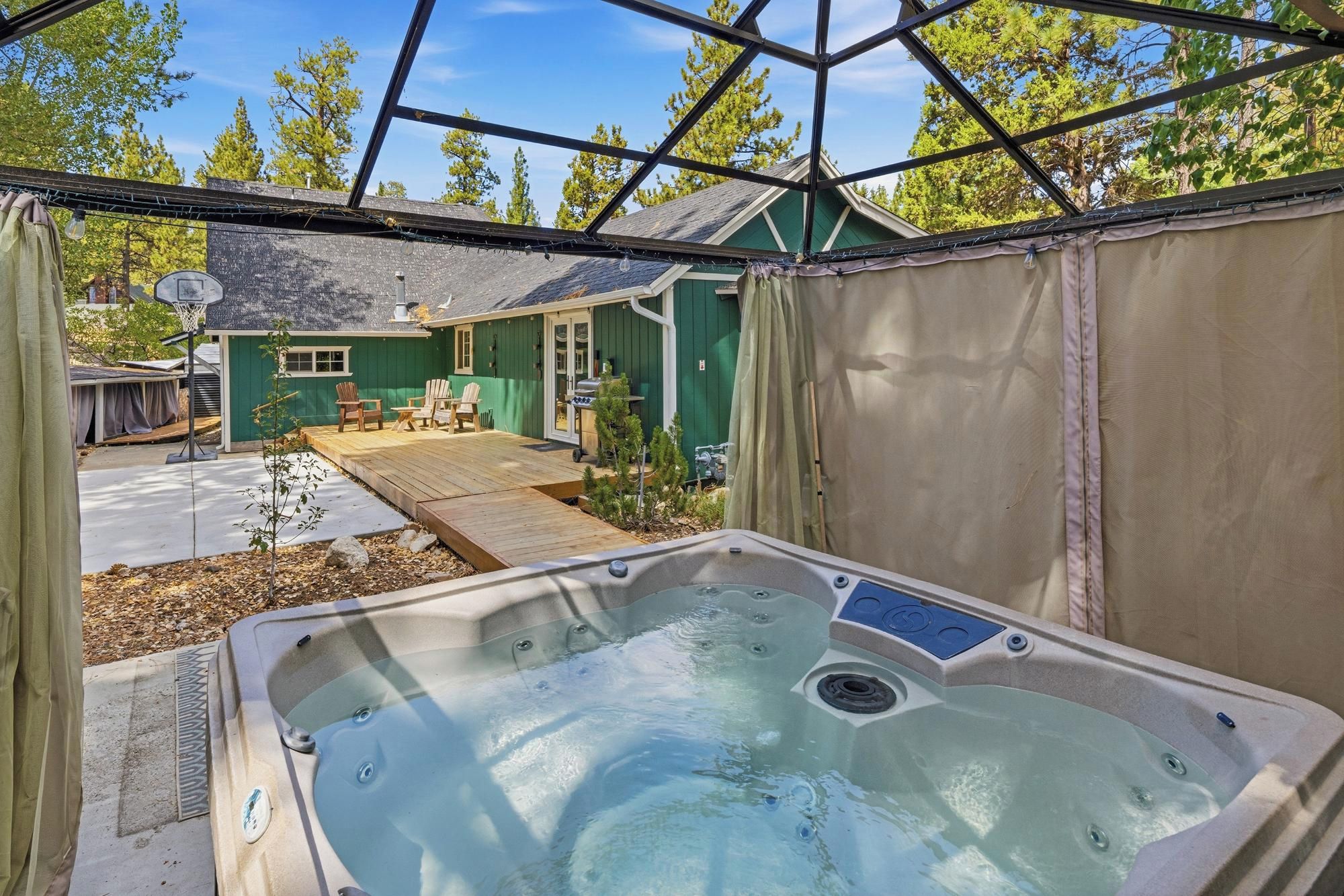 A hot tub in a backyard with a wooden deck, a green house, and a covered patio area.