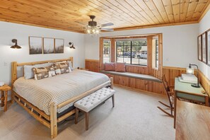 Ein Schlafzimmer mit Holzboden, einem Bett mit kariertem Überwurf, einer Bank, einem Schreibtisch mit Lampe und einem Fenster mit Blick ins Freie.