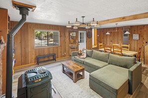 A cozy living room with a green sofa, wooden dining table, and a wood-burning stove. - HOT TUB! PET FRIENDLY! Basketball! Fire Pit! EV Charger! A/C! (Big Bear City)