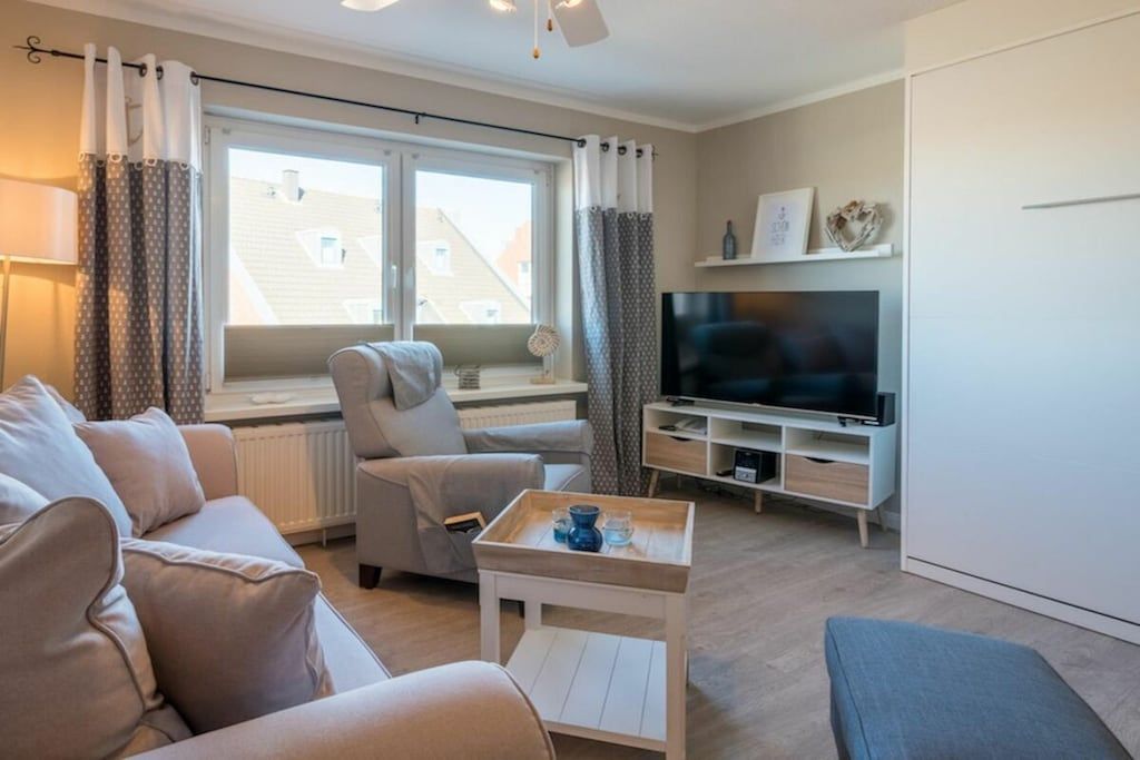 Ein modernes Wohnzimmer mit einer Couch, einem Sessel und einem Couchtisch. Ein Fernseher steht auf einem Schrank und ein Fenster ist mit Vorhängen versehen.