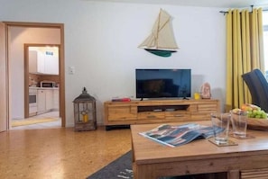 Ein Wohnzimmer mit Fernseher, Holzmöbeln, einer Segelboot-Dekoration und Blick in die Küche.